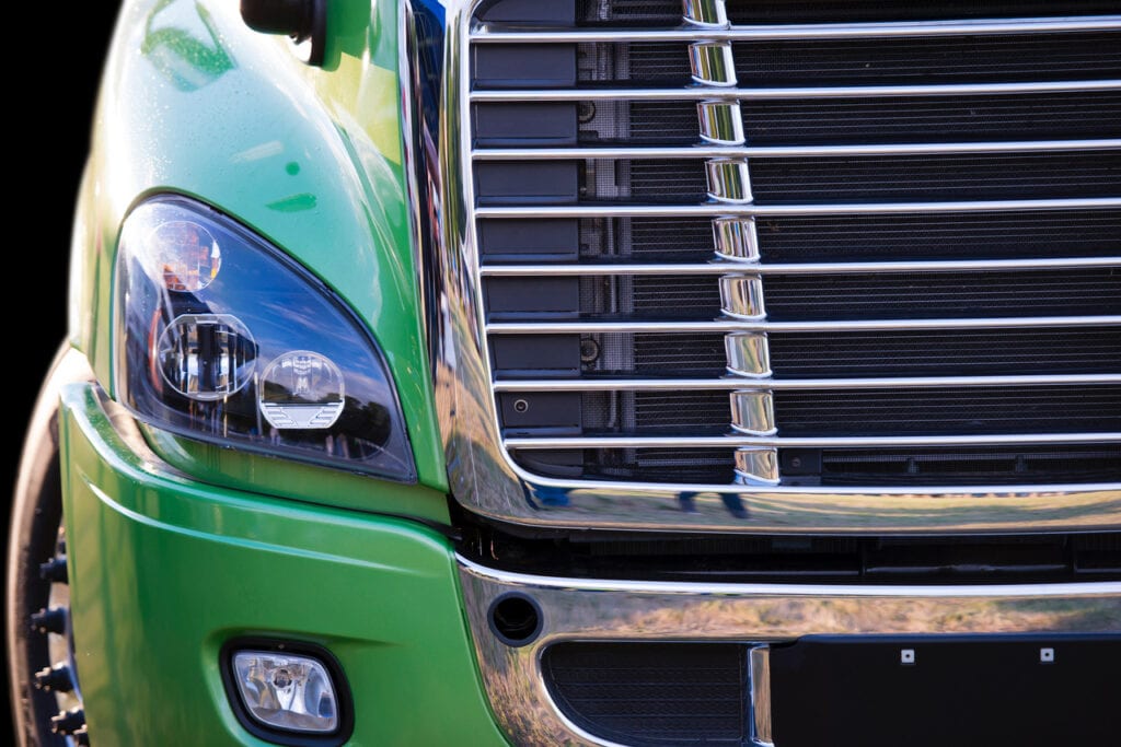 Sunlit stylish and comfortable green big rig semi truck of latest model of commercial long-distance transport with shiny chrome grille and efficient headlight in the parking lot waiting for cargo (Sunlit stylish and comfortable green big rig semi truck)